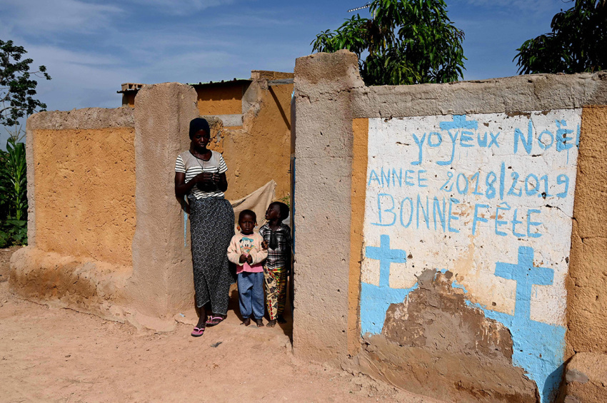 A woman and children stand at a fence with mural reading 'Merry Christmas happy celebrations' in the village of Yagma, north of Ouagadougou, on September 17, 2019. Burkina Faso, a poor country in West Africa, has been caught in a spiral of violence attributed to jihadist armed groups.