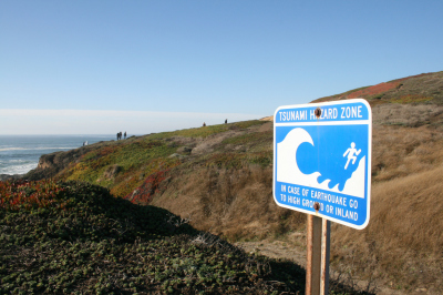 Bodega Bay, United States - November 14, 2015: Blue and white sign warning of a tsunami hazard zone along the San Andrea Fault in Bodega Bay, California, with hikers visible in the background.