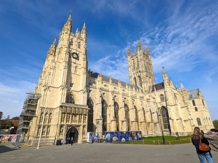 Visitors walk near the grand facade and towering spires of the historic Gothic Canterbury Cathedral in Canterbury, Kent, England.