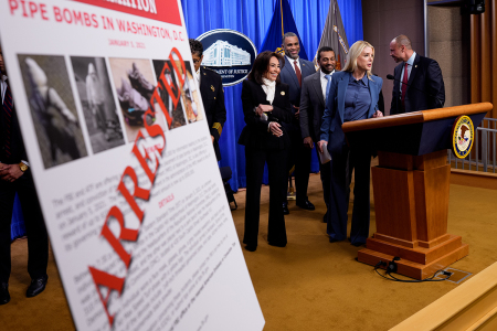 A poster reads "ARRESTED" in bold letters as Attorney General Pam Bondi, accompanied by U.S. Attorney for the District of Columbia Jeanine Pirro (L), FBI Director Kash Patel, (3rd-R) and FBI Deputy Director Dan Bongino (R), speaks at the conclusion of a news conference on an arrest of a suspect in the Jan. 6, 2021, pipe bomber at the Department of Justice on Dec. 4, 2025 in Washington, D.C. Federal agents have arrested a suspect they are charging with placing two pipe bombs, which never exploded, the night before the Jan. 6, 2021, U.S. Capitol attack.