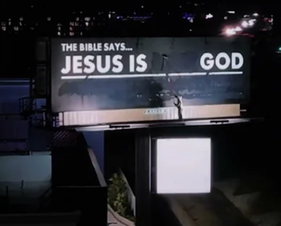 A screenshot of a video showing an unidentified man painting over a billboard in Los Angeles to read "The Bible says ... Jesus is ___ God."