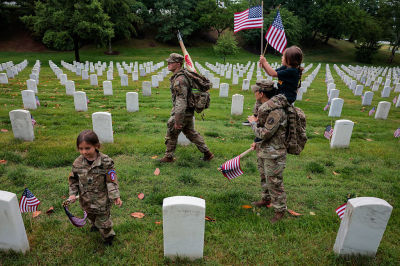 A member of the 3rd U.S. Infantry Regiment with her daughter on her shoulders places flags in front of each headstone at Arlington National Cemetery on May 21, 2026 in Arlington, Virginia. Before dawn soldiers begin the process of placing a flag in front of approximately 260,000 headstones ahead of Memorial Day.