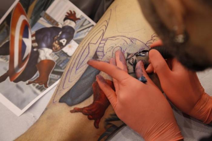 A tattoo artist from Virginia works on a man's leg with a Captain America design.