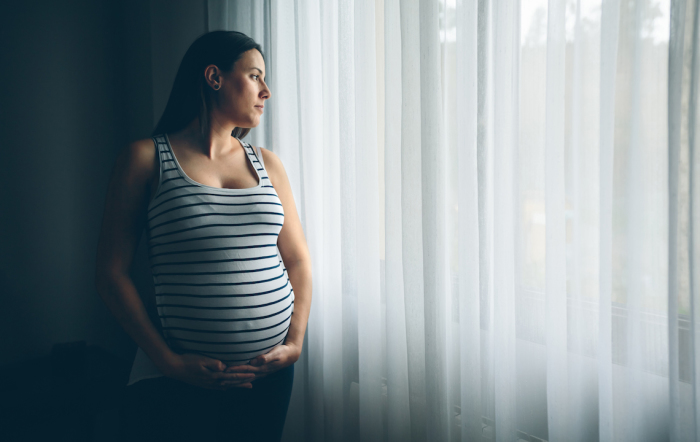 An expectant mother stares out the window as she holds her pregnant belly. 