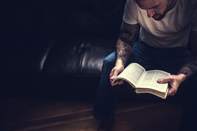 An overhead view of a man reading scripture in a dark room, window light illuminating the pages from above and behind him. Horizontal image with copy space.