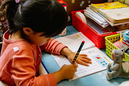 A student sits at her desk and writes on a piece of paper with a writing utensil.