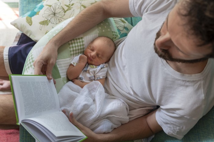 Father reading a book on the sofa with his baby daughter sleeping on his lap on the sofa.