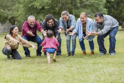 A multi-generation Hispanic family standing in the park together. A 16 month old baby girl is the center of attention. Her back is to the camera as she walks toward her family, all smiling with their arms open, reaching for her.