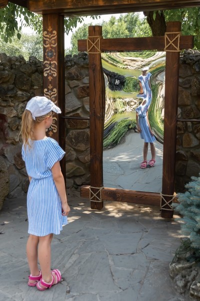 Little girl in front of distorting mirror. Funhouse mirror in a park.