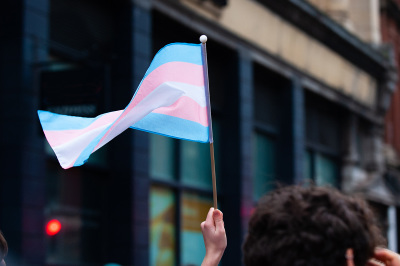 A hand holds up a small transgender pride flag. The blue and pink stripes represent the colors for a boy and girl, while the white stripe represents self-declared gender identities, such as transitioning, intersex, neutral and undefined gender.