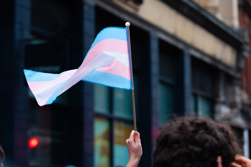 A hand holds up a small transgender pride flag. The blue and pink stripes represent the colors for a boy and girl, while the white stripe represents self-declared gender identities, such as transitioning, intersex, neutral and undefined gender.