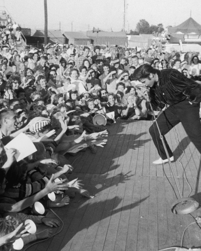 circa 1957: American singer and actor Elvis Presley (1935-1977) performing outdoors on a small stage to the adulation of a young crowd.