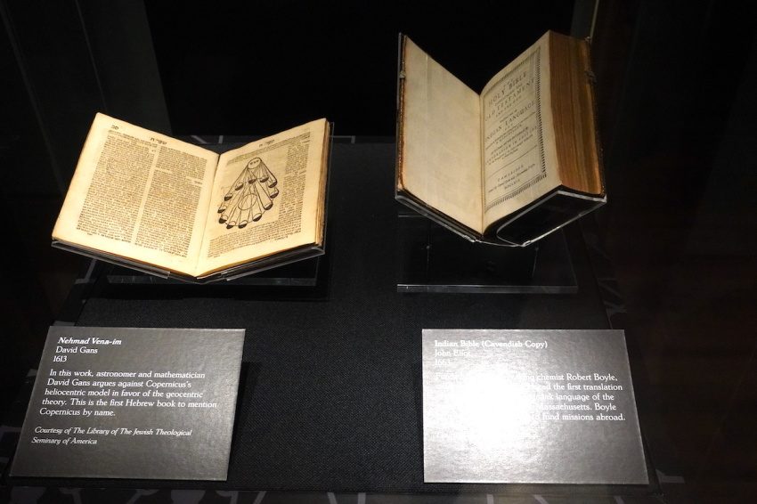 Pictured on the left is the Nehmad Vena-im by David Gans of 1613. In this work, astronomers and mathematician David Gans argues against Copernicus's heliocentric model in favor of geocentric theory. This is the first Hebrew book to mention Copernicus by name. The work is included in the Scripture and science exhibit as a courtesy of The Library of The Jewish Theological Seminary of America. On the right is pictured what is known as the Indian Bible by missionary John Eliot, who produced in its pages the first translation of the Bible in the Wôpanâak language of the local Native Americans of Massachusetts, courtesy of The Signatry.