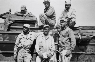 Six Black United States soldiers (Technician fifth grade LC Carter, Jr, Private John Bonner, Jr, Staff Sergeant Charles R Johnson (standing) Technician fifth grade AB Randle, Technician fifth grade Homer H. Gaines, and Private Willie Tellie) pose beside their amphibious vehicle, possibly a DUKW, location unspecified, Japan, 11th March 1945. The six soldiers has rescued an unspecified US Marine who was drowning in the surf at Iwo Jima, as their amphibian truck was swamped by heavy seas.