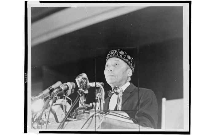 Elijah Muhammad stands behind microphones at a podium in 1964. 