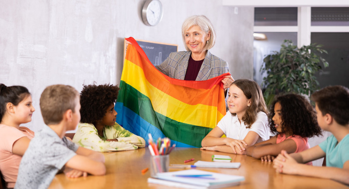 A teacher holds up a rainbow flag representing the LGBT movement in front of a classroom of students.