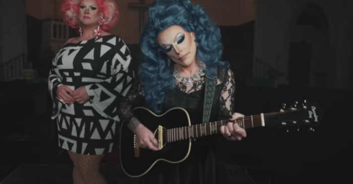 Drag performer Matthew Blake (left) and CCM singer Derek Webb wear makeup and women's clothing while standing in front of a cross for a music video. 