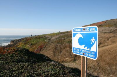 Bodega Bay, United States - November 14, 2015: Blue and white sign warning of a tsunami hazard zone along the San Andrea Fault in Bodega Bay, California, with hikers visible in the background.