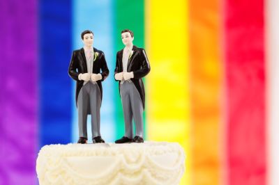 Two male groom figurine cake toppers sit atop a same-sex marriage wedding cake with a rainbow flag in background. 