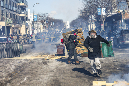 Protesters take cover behind overturned trash containers as a line of federal agents forms a barricade across a downtown street during clashes following the fatal shooting of an armed demonstrator earlier in the day, on Jan. 24, 2026, in Minneapolis, Minnesota. 