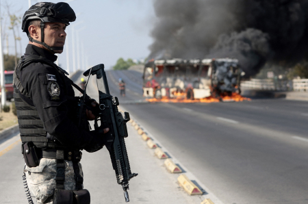 A member of the Prosecutor's Office stands guard near a burning bus at one of the main avenues after it was set on fire by organized crime groups in response to an operation in Jalisco to arrest a high-priority security target in Zapopan, state of Jalisco, Mexico, on Feb. 22, 2026. Armed civilians blocked several roads in the state of Jalisco, in western Mexico, following an operation by federal forces in the town of Tapalpa, local authorities reported. Jalisco, which will host four matches of the upcoming 2026 World Cup, is home to the powerful Jalisco New Generation Cartel (CJNG), and has been rocked by several episodes of violence in recent years. 