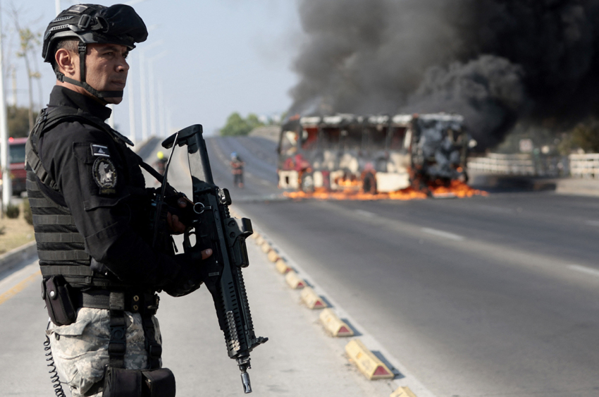 A member of the Prosecutor's Office stands guard near a burning bus at one of the main avenues after it was set on fire by organized crime groups in response to an operation in Jalisco to arrest a high-priority security target in Zapopan, state of Jalisco, Mexico, on Feb. 22, 2026. Armed civilians blocked several roads in the state of Jalisco, in western Mexico, following an operation by federal forces in the town of Tapalpa, local authorities reported. Jalisco, which will host four matches of the upcoming 2026 World Cup, is home to the powerful Jalisco New Generation Cartel (CJNG), and has been rocked by several episodes of violence in recent years. 