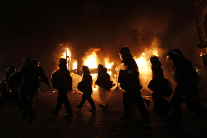 Police officers wearing riot gear walk past a burning building in Tottenham, north London August 7, 2011. Crowds attacked riot police and set two squad cars alight in north London on Saturday following a protest at the fatal shooting of a man by armed officers earlier in the week.