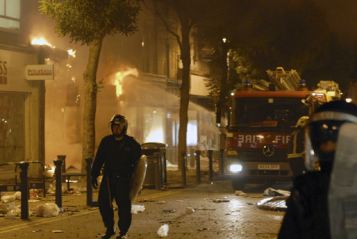 A police officer stands guard as firefighters work to extinguish the flames of a blazing store in Woolwich, southeast London, August 9, 2011. Rioting and looting spread across and beyond London on Monday as hooded youths set fire to cars and buildings, smashed shop windows and hurled bottles and stones at police in a third night of violence in Britain's worst unrest in decades.