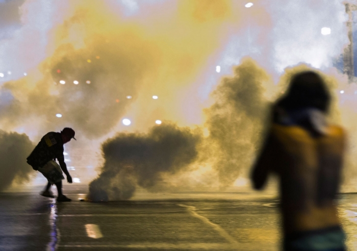 A protester reaches down to throw back a smoke canister as police clear a street after the passing of a midnight curfew meant to stem ongoing demonstrations in reaction to the shooting of Michael Brown in Ferguson, Missouri August 17, 2014. The group of protesters angry at the shooting death of Brown, a black teenager, by a white police officer remained on the streets of Ferguson, Missouri, early on Sunday minutes past the declared curfew, as police gathered nearby in a tense standoff.