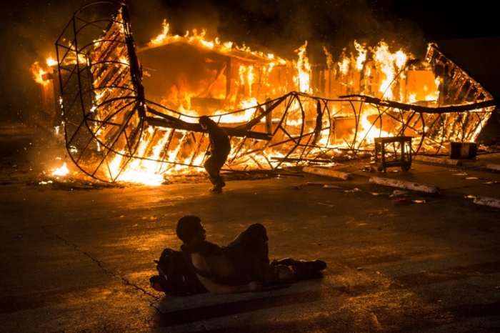 A resident, lying shirtless, keeps warm as another approaches the blazing skeleton of Juanita's Fashions R Boutique after it was burned to the ground in Ferguson, Missouri, early morning November 25, 2014. Gunshots rang out and buildings burned in a Midwestern suburb after a grand jury decided on Monday not to indict a white police officer over the fatal August shooting of an unarmed black teenager, sparking a fresh wave of racially tinged violence. At least a dozen buildings were set on fire in Ferguson, Missouri, most of them left destroyed.