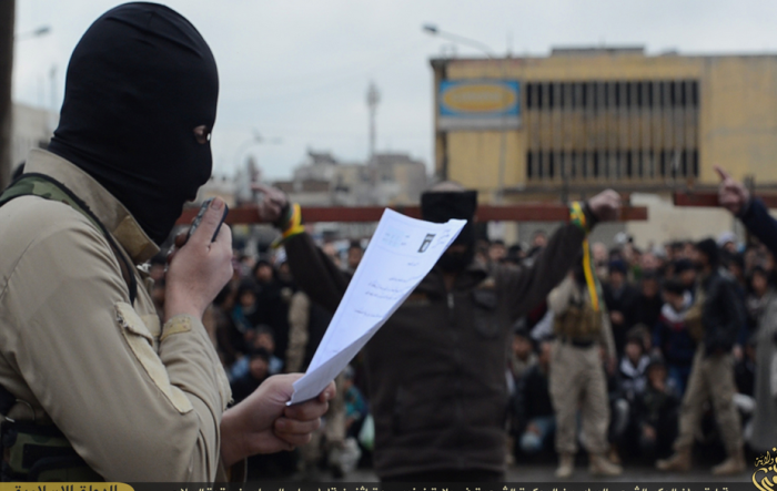 A masked ISIS militant reads the charges facing the two men tied to a cross, who were later shot in the back of the head for banditry, Mosul, Iraq.