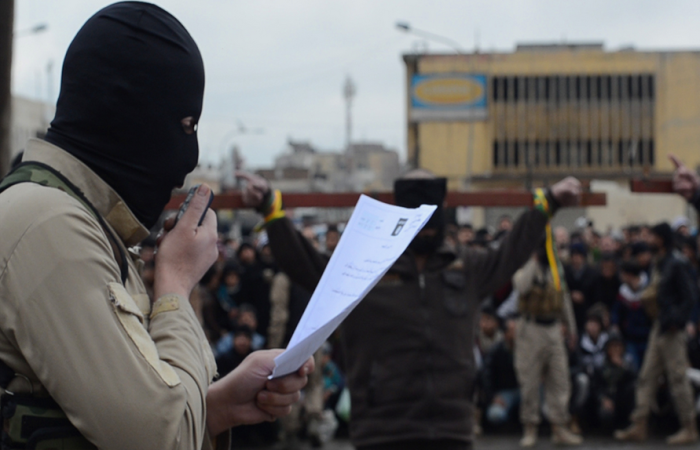A masked ISIS militant reads the charges facing the two men tied to a cross, who were later shot in the back of the head for banditry, Mosul, Iraq.