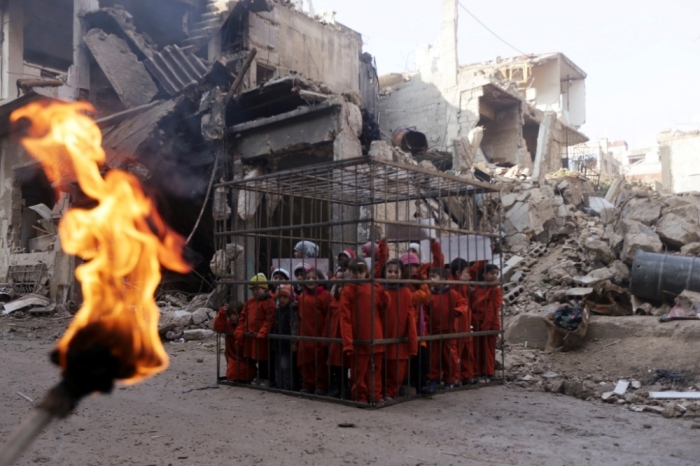 An activist (not pictured) holds a burning torch near children carrying banners inside a cage during a protest against forces loyal to Syria's President Bashar al-Assad in Douma, eastern Al-Ghouta, near Damascus, February 15, 2015. The protest, which made children wear orange suits depicting victims of the Islamic State, calls to compare forces loyal to Syria's president Bashar al-Assad to Islamic State forces, and to draw attention to residents living under siege and dying from strikes by forces loyal to Syria's president Bashar Al-Assad, activists said.