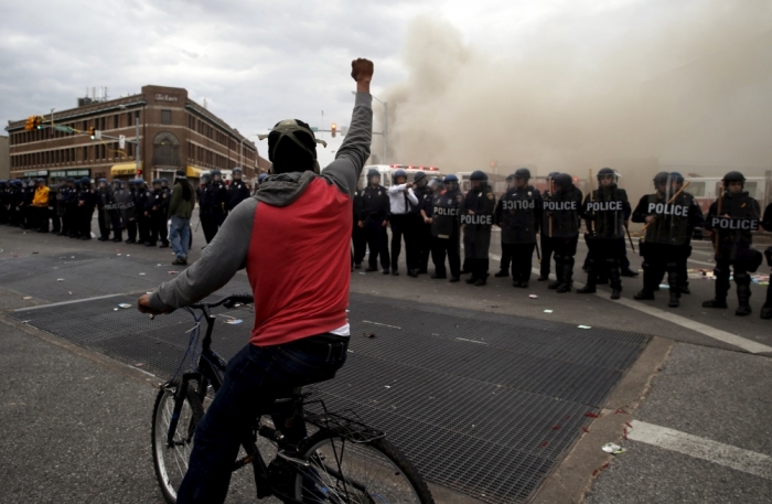 A protestor on a bicycle thrusts his fist in the air next to a line of police, in front of a burning CVS drug store, during clashes in Baltimore, Maryland, April 27, 2015. Maryland Governor Larry Hogan declared a state of emergency and activated the National Guard to address the violence in Baltimore, his office said on Monday. Several Baltimore police officers were injured on Monday in violent clashes with young people after the funeral of a black man, Freddie Gray, who died in police custody, and local law enforcement warned of a threat by gangs.