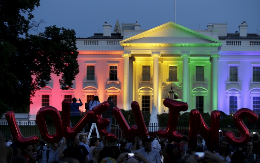 People stand in front of the rainbow-colored illuminated White House with balloons that say 
