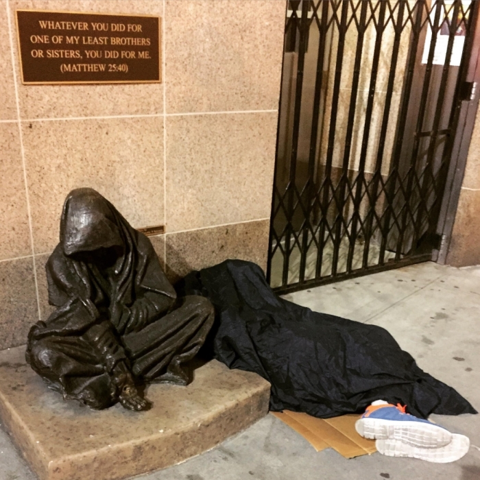 A homeless man sleeps next to a bronze statue inspired by Matthew 25:40 named 'Whatsoever You Do' created by Canadian sculptor Timothy Schmalz.
