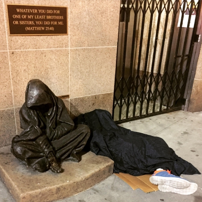 A homeless man sleeps next to a bronze statue inspired by Matthew 25:40 named "Whatsoever You Do" created by Canadian sculptor Timothy Schmalz.