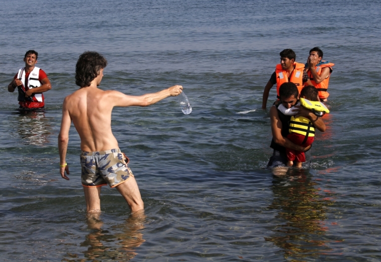 A tourist offers water to Iranian migrants as they arrive by paddling an engineless dinghy from the Turkish coast (seen in the background) at a beach on the Greek island of Kos August 15, 2015. United Nations refugee agency called on Greece to take control of the 'total chaos' on Mediterranean islands, where thousands of migrants have landed. About 124,000 have arrived this year by sea, many via Turkey, according to Vincent Cochetel, UNHCR director for Europe. .