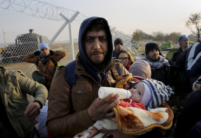 A Syrian refugee feeds his child in front of a fence at the Greek-Macedonian border, while waiting to cross over, near to the village of Idomeni, Greece, December 5, 2015.