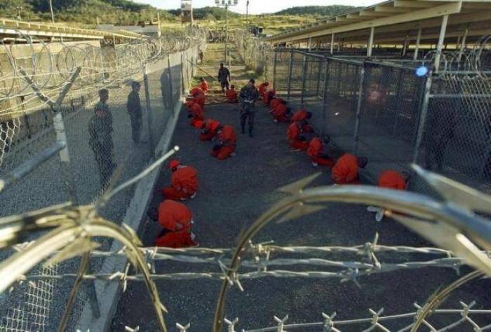 Detainees in orange jumpsuits sit in a holding area under the watchful eyes of military police during in-processing to the temporary detention facility at Camp X-Ray of Naval Base Guantanamo Bay in this January 11, 2002 file photograph.